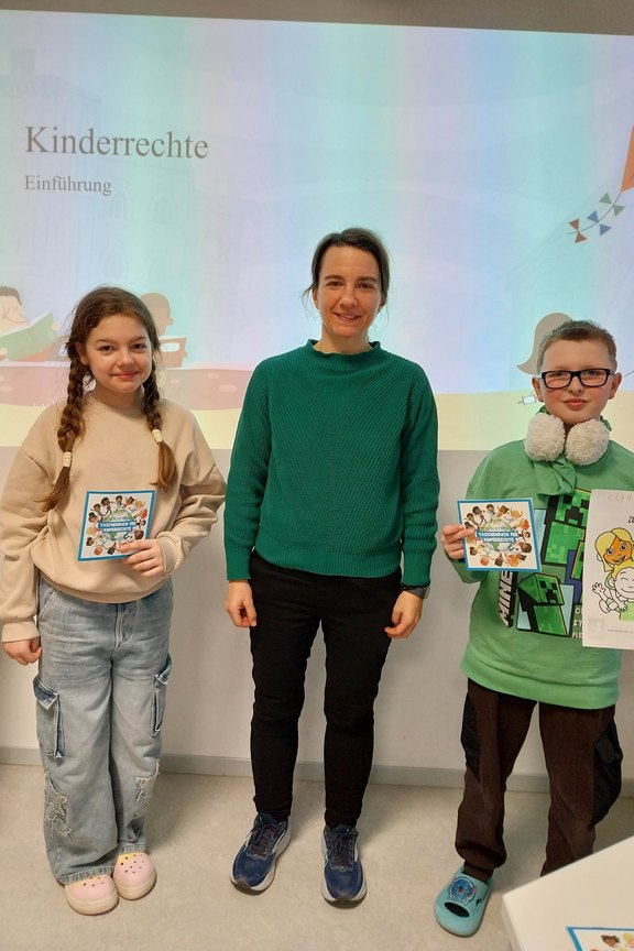 Aniko Ligeti Das Bild zeigt zwei Kinder, die ein ausgemaltes Bild zeigen. In deren Mitte steht eine Frau.