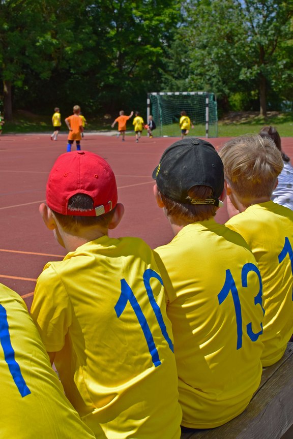 Das Bild zeigt Kinder in Trikots nebeneinander auf einer Bank sitzend. Im Hintergrund ist ein Fußballspiel zu sehen.  